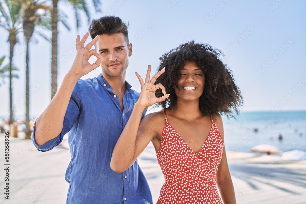 Naklejka premium Young interracial couple outdoors on a sunny day smiling positive doing ok sign with hand and fingers. successful expression.