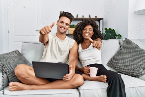 Young interracial couple using laptop at home sitting on the sofa approving doing positive gesture with hand, thumbs up smiling and happy for success. winner gesture.