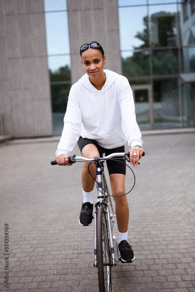 Young african american woman in cycling shorts and hoodie looking at camera while biking on street 