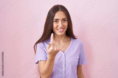 Young hispanic woman with long hair standing over pink background beckoning come here gesture with hand inviting welcoming happy and smiling
