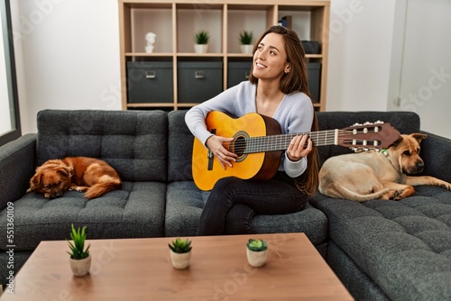 Young hispanic woman playing classical guitar sitting on sofa with dogs at home