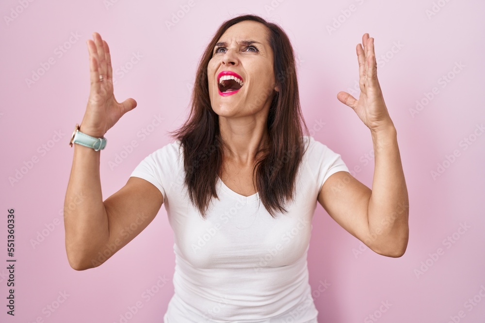 Middle age brunette woman standing over pink background crazy and mad shouting and yelling with aggressive expression and arms raised. frustration concept. Middle age brunette woman standing over pink background crazy and mad shouting and yelling with aggressive expression and arms raised. frustration concept.