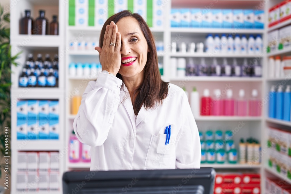 Middle age brunette woman working at pharmacy drugstore covering one eye with hand, confident smile on face and surprise emotion.
