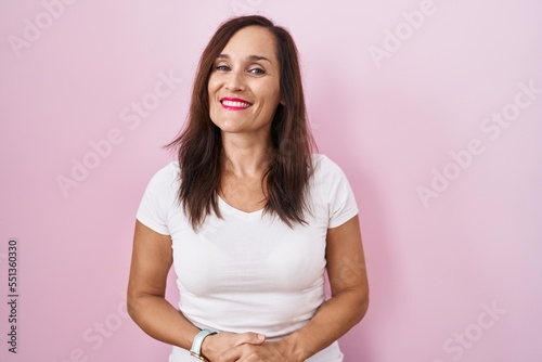 Middle age brunette woman standing over pink background with hands together and crossed fingers smiling relaxed and cheerful. success and optimistic