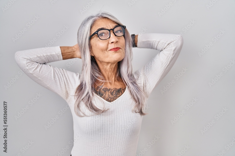 Middle age woman with grey hair standing over white background relaxing and stretching, arms and hands behind head and neck smiling happy Middle age woman with grey hair standing over white background relaxing and stretching, arms and hands behind head and neck smiling happy