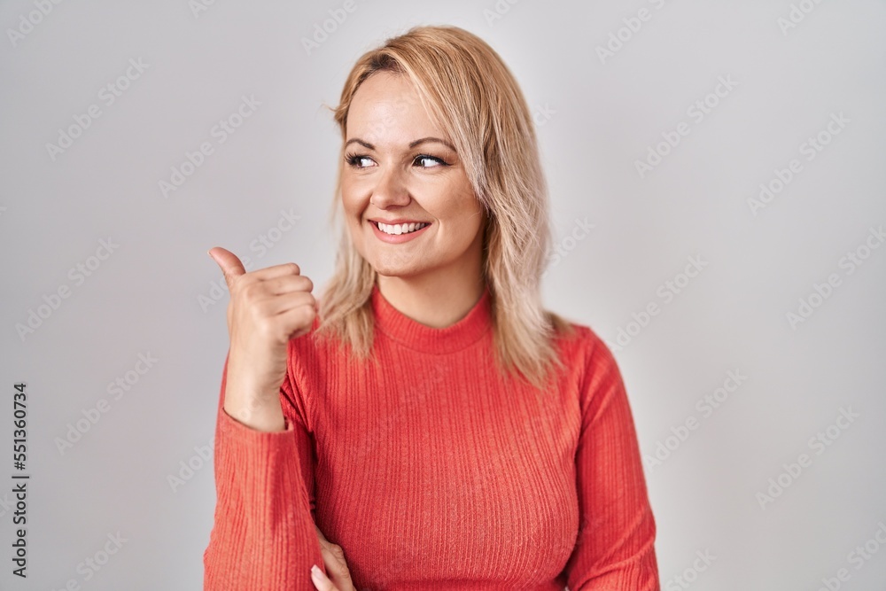 Blonde woman standing over isolated background smiling with happy face looking and pointing to the side with thumb up. Blonde woman standing over isolated background smiling with happy face looking and pointing to the side with thumb up.