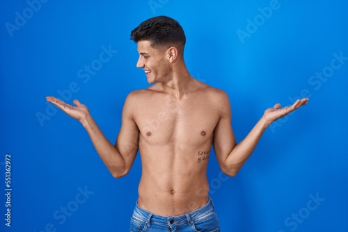 Young hispanic man standing shirtless over blue background smiling showing both hands open palms, presenting and advertising comparison and balance