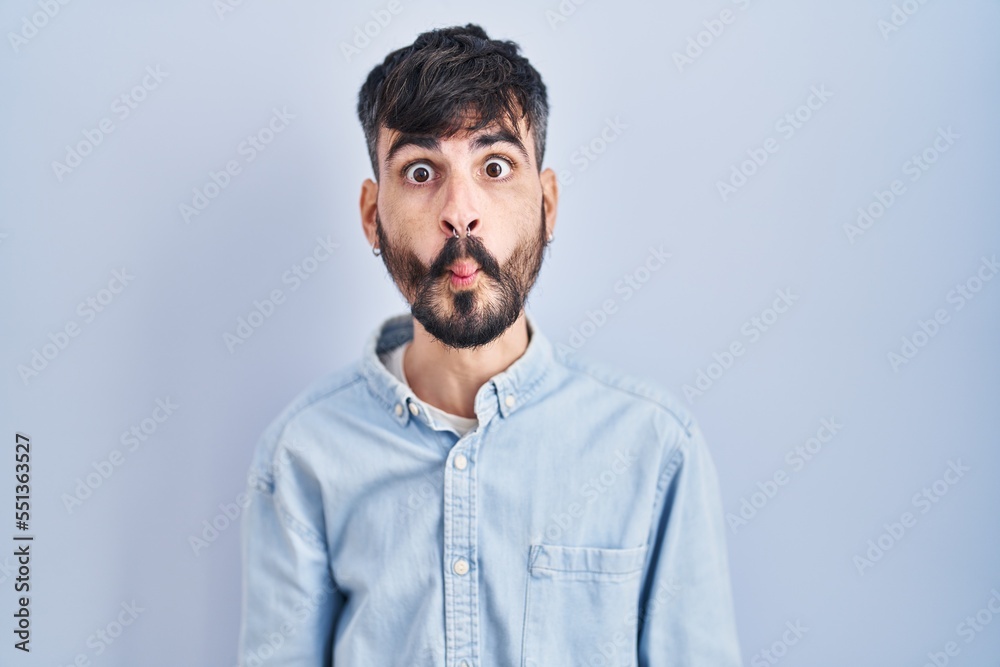 Young hispanic man with beard standing over blue background making fish face with lips, crazy and comical gesture. funny expression. Young hispanic man with beard standing over blue background making fish face with lips, crazy and comical gesture. funny expression.