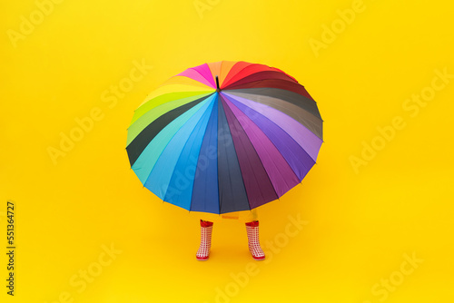 A multicolored rainbow umbrella with legs in red rubber boots on a yellow isolated background. Rainy spring, autumn weather. Legs peek out from under the umbrella.