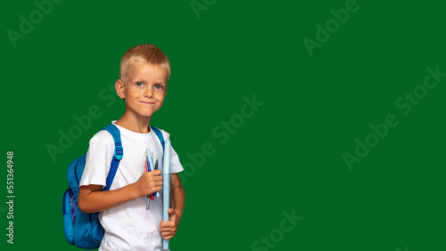 Boy with backpack is smiling, with book and markers in his hands. Green background with space for text. Selective focus. Picture for articles about children, school, education.