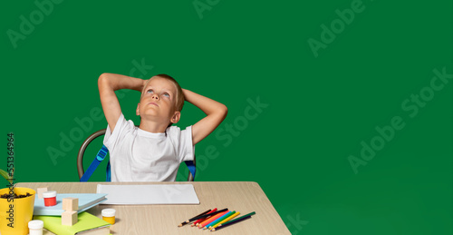 Boy puts his hands behind his head and looks up. There are pencils, book, cubes, paints on table. Training, coercion, discontent, disobedience, education, denial. Selective focus.