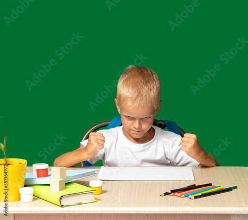 Disgruntled boy sits at table with his fists clenched. There are pencils, book, cubes, paints on table. Training, coercion, discontent, disobedience, upbringing. Selective focus.
