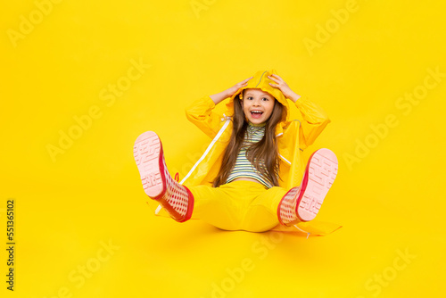 A child in a raincoat, sitting with his legs up in red boots. A beautiful little girl is smiling and sitting on a yellow isolated background. Rainy spring, autumn weather. © Юлия Дьякова