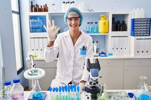 Brunette woman working at scientist laboratory showing and pointing up with fingers number five while smiling confident and happy.