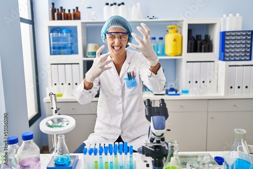 Brunette woman working at scientist laboratory shouting frustrated with rage  hands trying to strangle  yelling mad