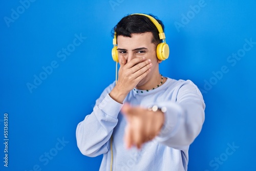 Non binary person listening to music using headphones laughing at you, pointing finger to the camera with hand over mouth, shame expression