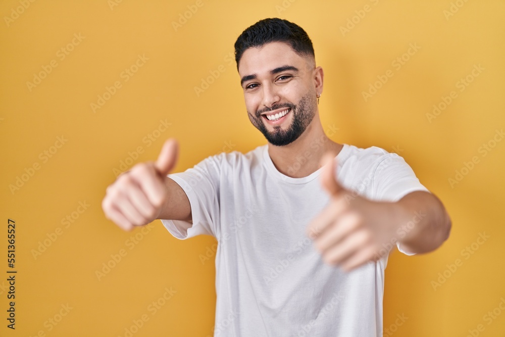Young handsome man wearing casual t shirt over yellow background approving doing positive gesture with hand, thumbs up smiling and happy for success. winner gesture. Young handsome man wearing casual t shirt over yellow background approving doing positive gesture with hand, thumbs up smiling and happy for success. winner gesture.