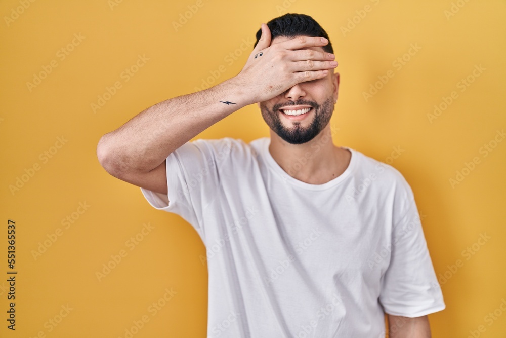 Young handsome man wearing casual t shirt over yellow background smiling and laughing with hand on face covering eyes for surprise. blind concept. Young handsome man wearing casual t shirt over yellow background smiling and laughing with hand on face covering eyes for surprise. blind concept.