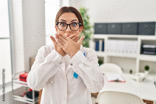 Young brunette doctor woman wearing stethoscope at the clinic shocked covering mouth with hands for mistake. secret concept.