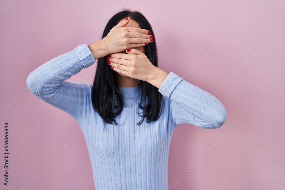 Hispanic woman standing over pink background covering eyes and mouth with hands, surprised and shocked. hiding emotion Hispanic woman standing over pink background covering eyes and mouth with hands, surprised and shocked. hiding emotion