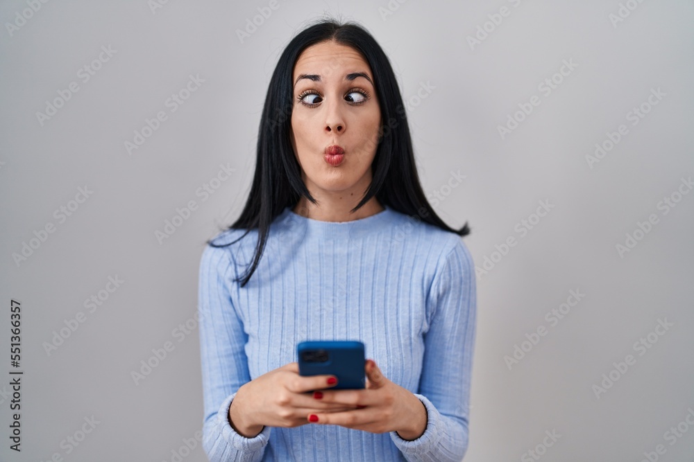 Hispanic woman using smartphone making fish face with mouth and squinting eyes, crazy and comical. Hispanic woman using smartphone making fish face with mouth and squinting eyes, crazy and comical.