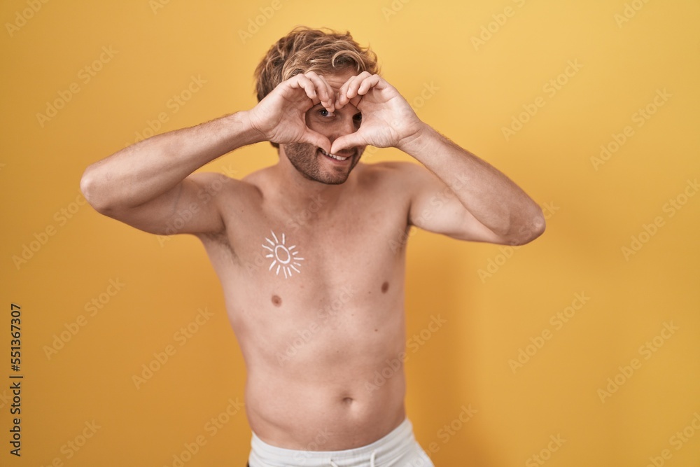 Fototapeta premium Caucasian man standing shirtless wearing sun screen doing heart shape with hand and fingers smiling looking through sign