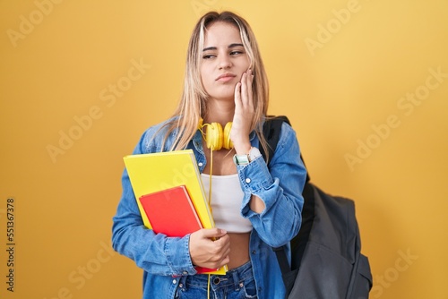 Young blonde woman wearing student backpack and holding books touching mouth with hand with painful expression because of toothache or dental illness on teeth. dentist concept.