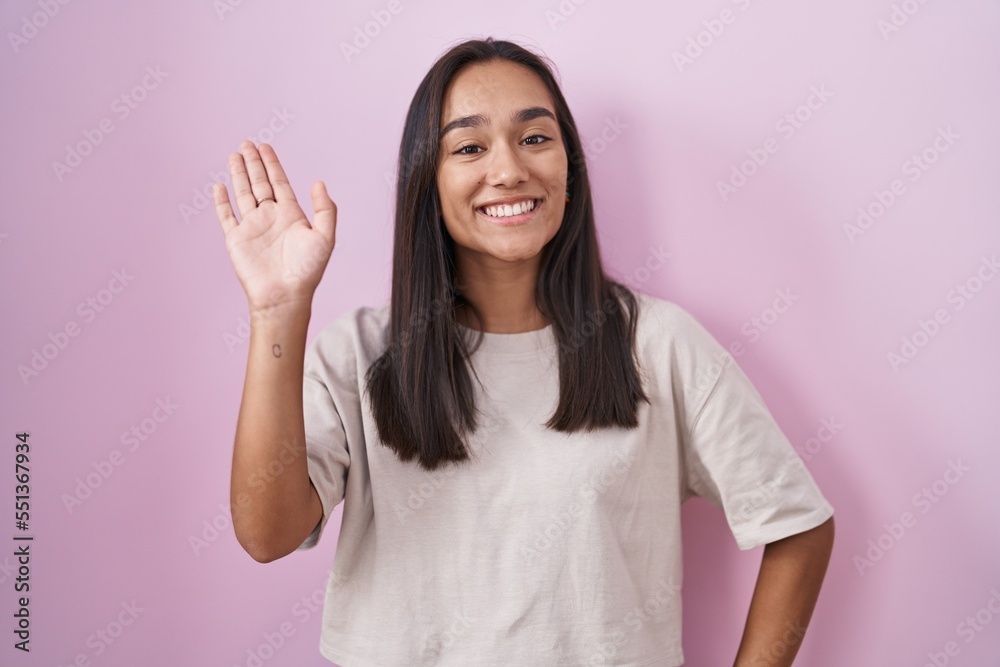 Young hispanic woman standing over pink background waiving saying hello happy and smiling, friendly welcome gesture Young hispanic woman standing over pink background waiving saying hello happy and smiling, friendly welcome gesture