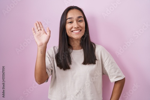 Young hispanic woman standing over pink background waiving saying hello happy and smiling, friendly welcome gesture