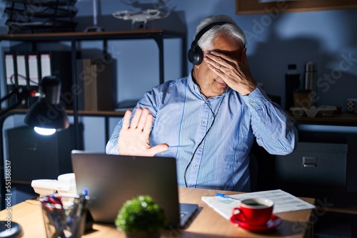 Hispanic senior man wearing call center agent headset at night covering eyes with hands and doing stop gesture with sad and fear expression. embarrassed and negative concept.