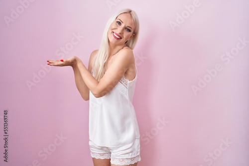 Caucasian woman wearing pajama wearing pink background pointing aside with hands open palms showing copy space, presenting advertisement smiling excited happy