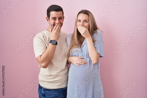 Young couple expecting a baby standing over pink background laughing and embarrassed giggle covering mouth with hands, gossip and scandal concept