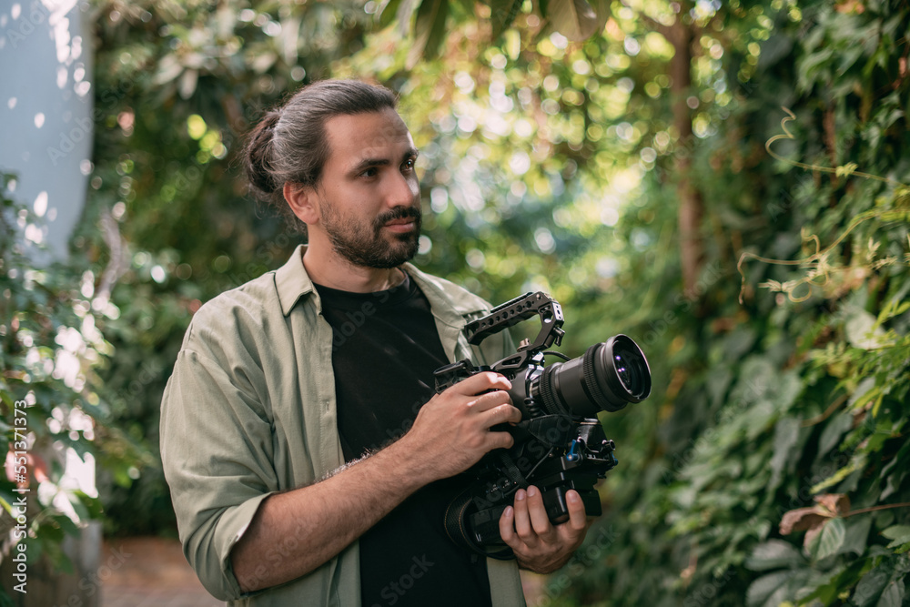 Fototapeta premium A young male cameraman with a professional movie camera in his hands in a green garden.