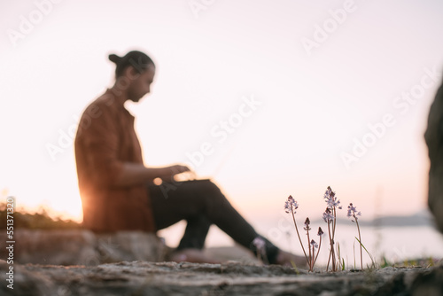 A young Caucasian man remotely works with a laptop in a garden on a mountain overlooking the sea and sunset.