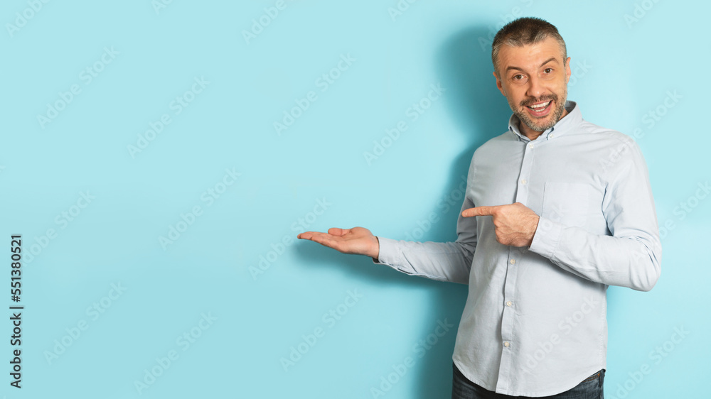 Excited caucasian mature man pointing finger at open palm, posing on blue background, panorama with free space Excited caucasian mature man pointing finger at open palm, posing on blue background, panorama with free space