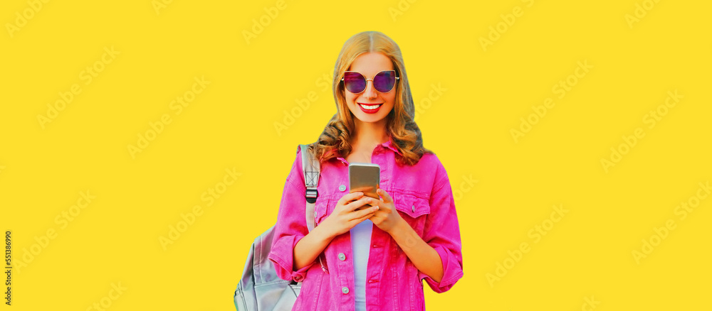 Portrait of stylish happy smiling young woman with smartphone wearing pink jacket, backpack and sunglasses on yellow background