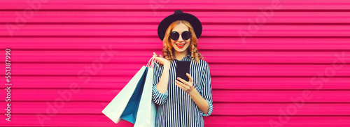 Portrait of happy smiling woman with shopping bags and smartphone wearing striped shirt, black round hat on pink background © rohappy