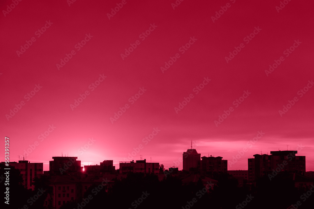 Silhouette of city at sunset. Sun is rising over tall buildings. Dramatic sky in sun rays. Urban landscape at dawn. Copy space. Color of the year 2023 - Viva Magenta