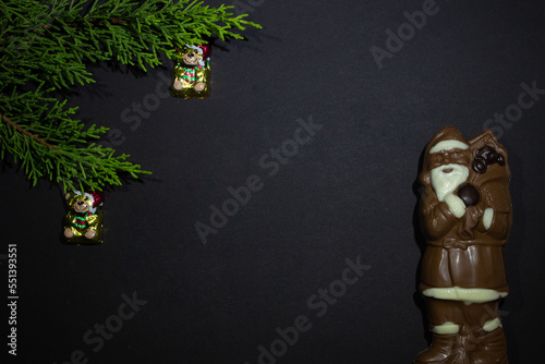 the photo of chocolate Santa Claus made of dark and white chocolate on black background and green fir branch and Christmas decorations bear