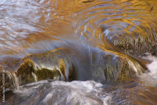 Das Wasser in einem Bach fließt über eine kleine Schwelle, die mit Moos bewachsen ist. Die Langzeitaufnahme verwischt die Konturen des Wassers, das dadurch wie eine Folie wirkt. Goldenes Herbstlaub.