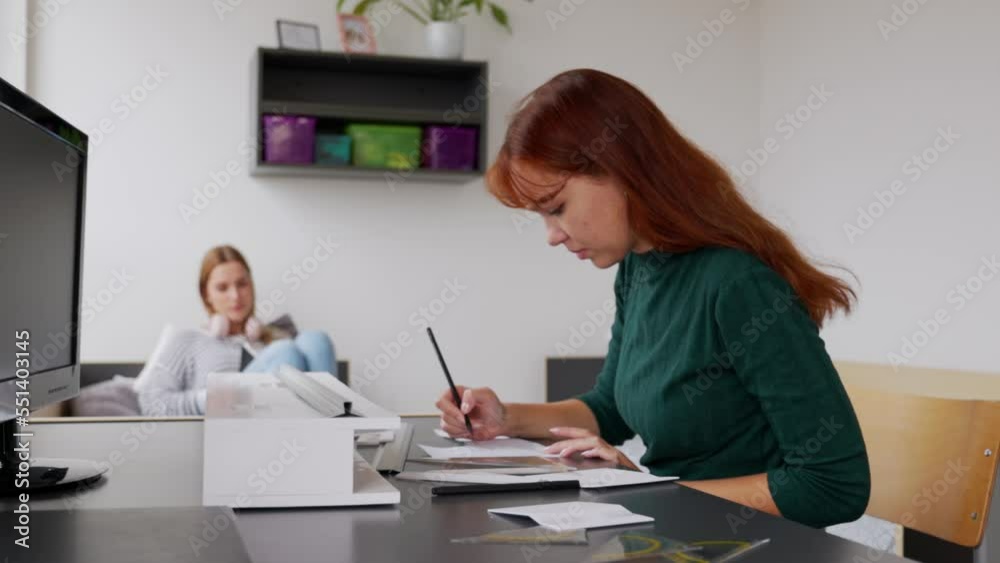 Two young women in their dorm room, the one with red hair working on her homework and the other laying on the bed reading a book Two young women in their dorm room, the one with red hair working on her homework and the other laying on the bed reading a book