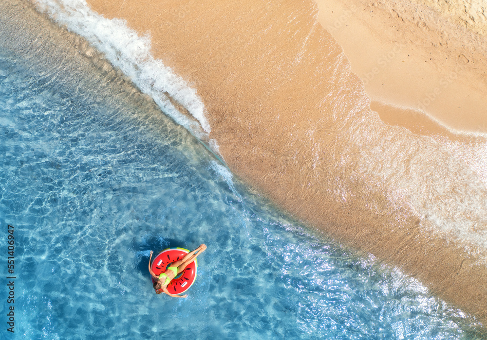 Aerial view of a young woman swimming with red swim ring in blue sea with waves at sunset in summer. Tropical landscape with girl, clear water, sandy beach. Top view. Vacation. Sardinia island, Italy	
