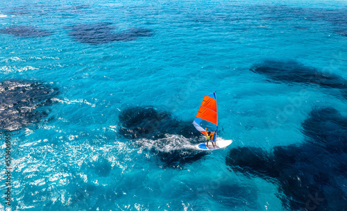 Aerial view of windsurfer on blue sea at sunny summer day. Windsurfing. Extreme sport and vacation. Top view of man on windsurfer board, waves, clear azure water in Sardinia, Italy. Tropical landscape