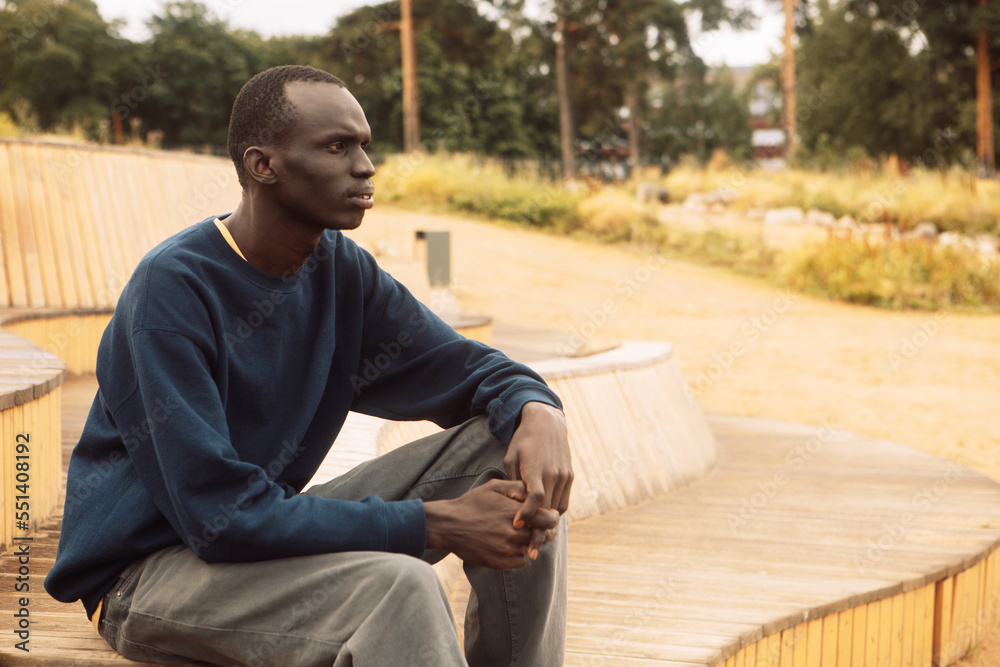 Obraz premium A dark-skinned young man sits on a wooden bench in the park and listens to music.