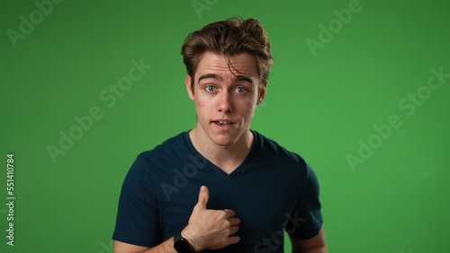 Confused young man 20s posing isolated on green screen background in studio. People lifestyle concept. Pointing fingers himself ask say who me waving and smiling