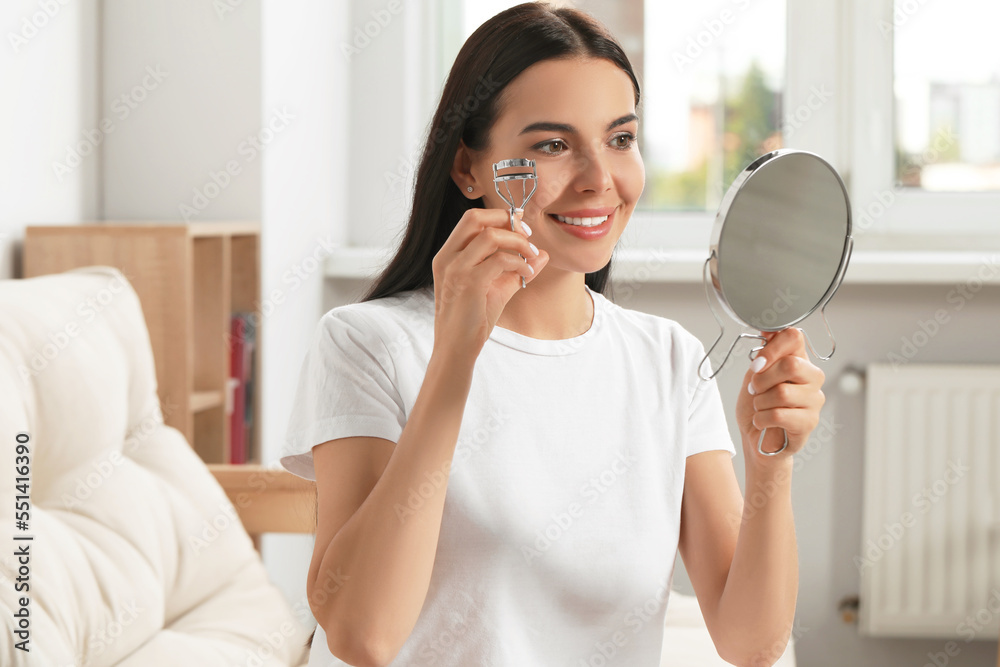 Beautiful young woman with mirror using eyelash curler indoors Beautiful young woman with mirror using eyelash curler indoors