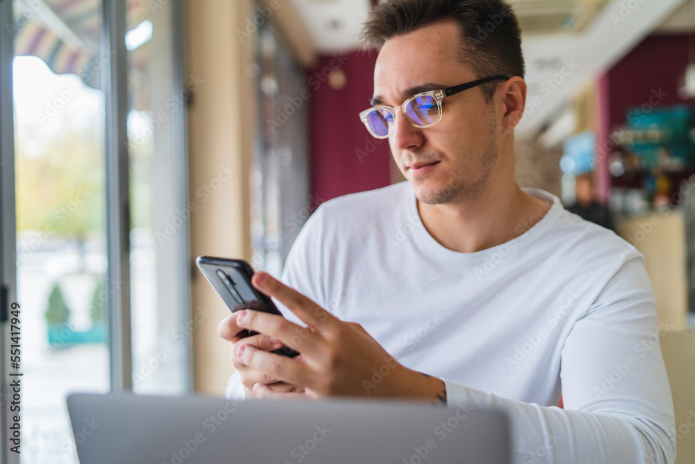 A young businessman in cafe using his laptop mobile phone and wireless earbuds while working on his project business on his laptop and talking on the phone A young businessman in cafe using his laptop mobile phone and wireless earbuds while working on his project business on his laptop and talking on the phone