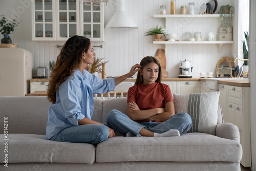 Kind caring woman straightens hair of girl of school age sits on sofa and not wanting to spend time with mother. Aggrieved introverted daughter sits with arms crossed because of resentment towards mom