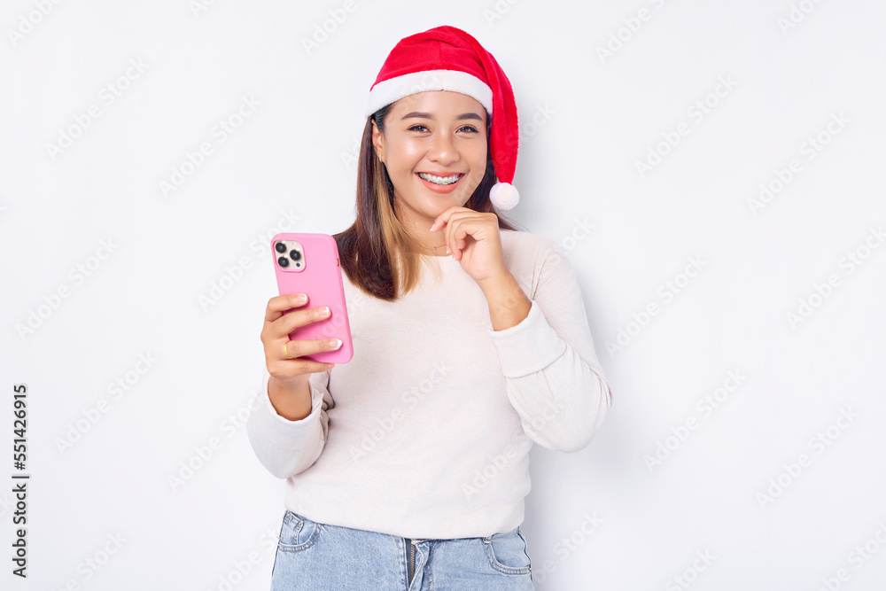 Smiling young Asian woman in a Christmas hat holding a mobile phone and touching her chin isolated over white background. celebration Christmas holiday and New Year concept Smiling young Asian woman in a Christmas hat holding a mobile phone and touching her chin isolated over white background. celebration Christmas holiday and New Year concept
