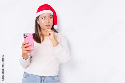 Pensive young Asian woman in a Christmas hat holding a mobile phone and  touching her chin isolated over white background